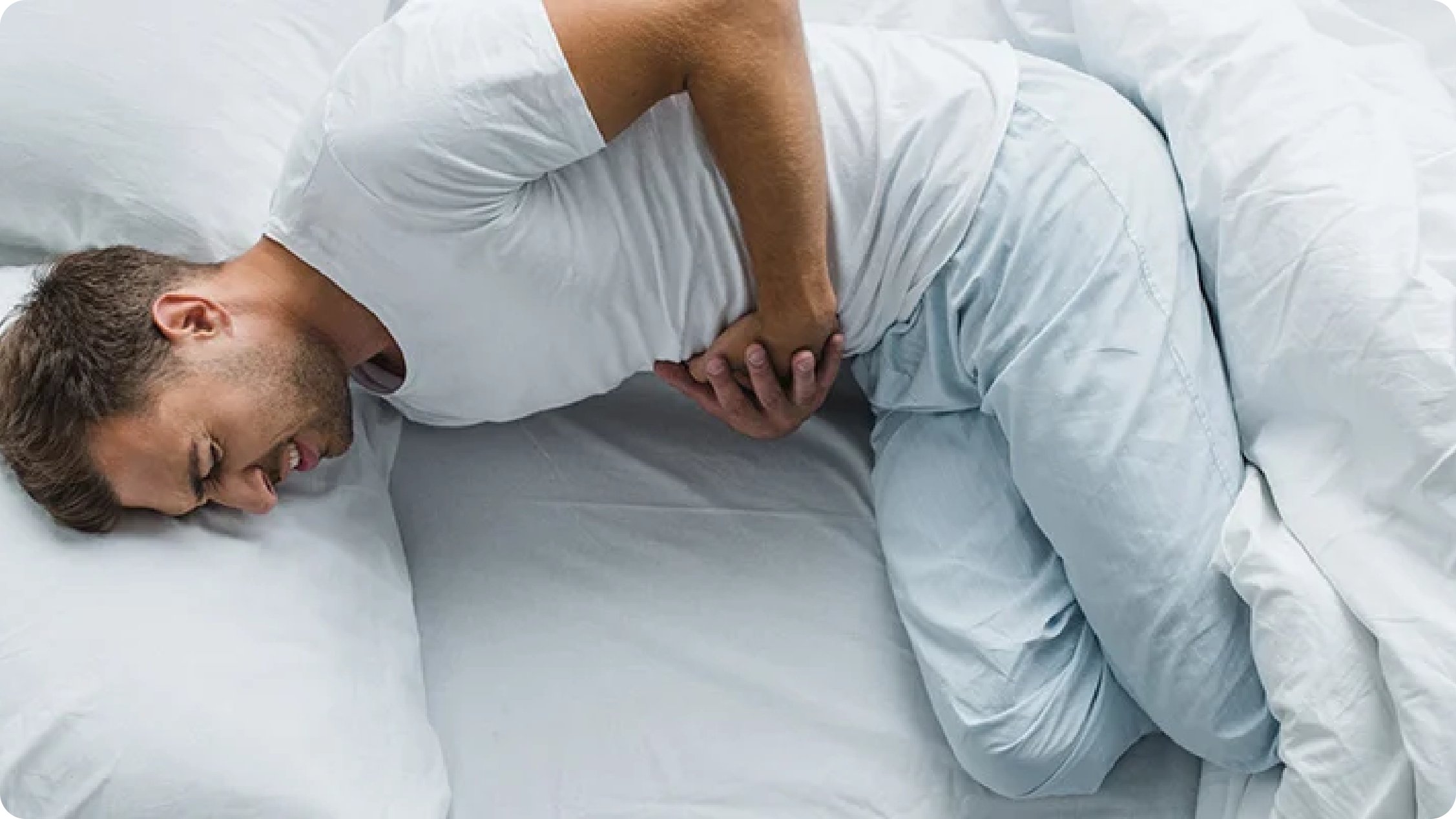 Ragazzo con maglia e pantaloni bianchi disteso su un letto con le mani sulla pancia e espressione dolorante