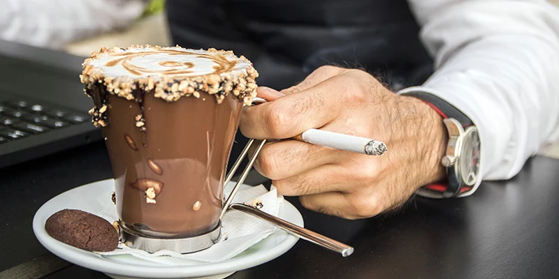 Uomo seduto al PC con un dettaglio della mano con una sigaretta e un bicchiere di cioccolata