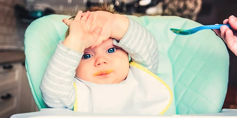 Bambino seduto su un seggiolone con le mani sulla fronte che sembra voler rifiutare la pappa