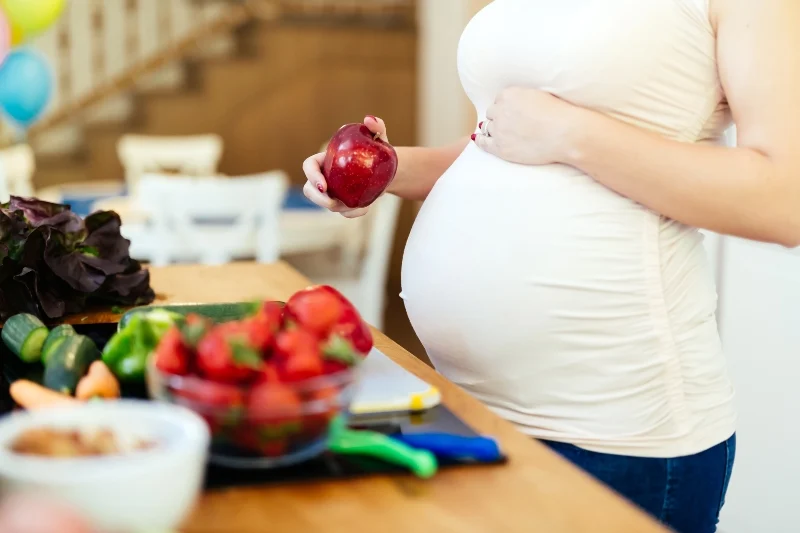 Donna in gravidanza con una mano sulla pancia, accanto ad un tavolo su cui posano cibi sani (peperoni, zucchine, carote, insalata), che ha in mano una mela