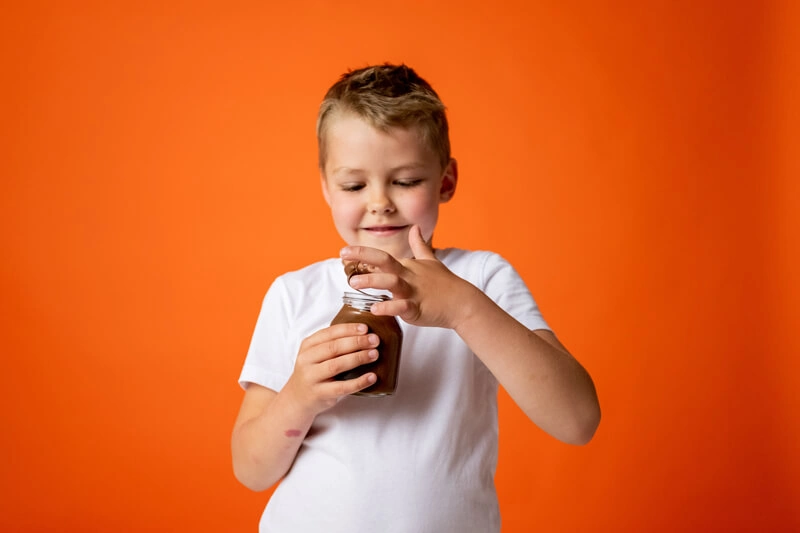 Bambino su sfondo arancione con un vasetto di marmellata in mano, in proconto di assaggiarla con le dita